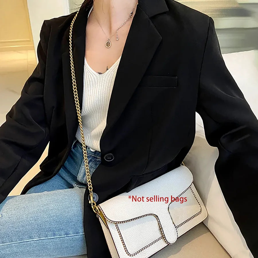 A woman models a black blazer and white top, holding a white quilted handbag with a gold chain strap.