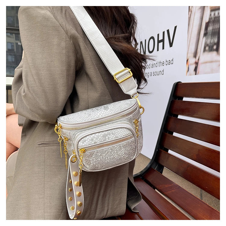 A woman wearing a beige blazer holds a silver and gold rivet waist bag crossbody against a light gray background.