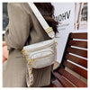 A woman wearing a beige blazer holds a silver and gold rivet waist bag crossbody against a light gray background.