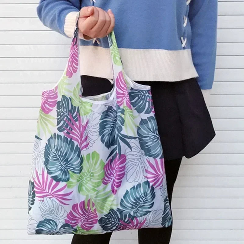 A person holds a white tote bag with a floral print featuring green and pink leaves against a white background.