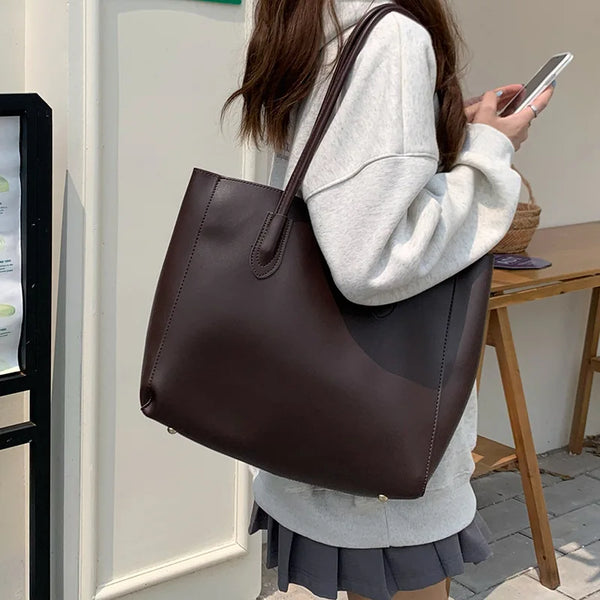 A woman holds a dark brown leather tote bag against a light background.