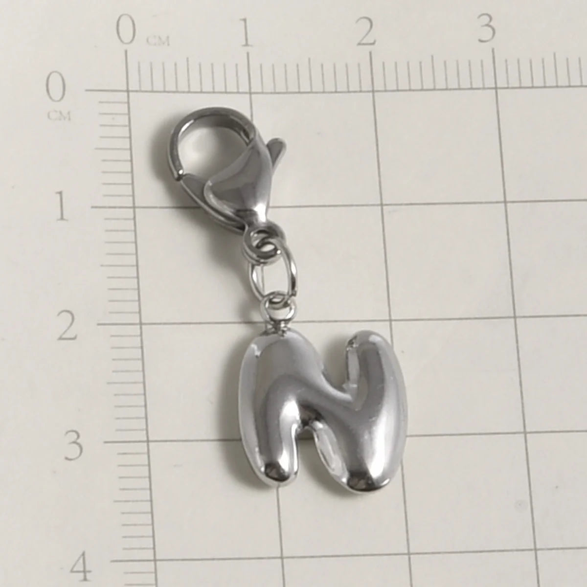 Silver stainless steel alphabet letter charm with a lobster clasp placed on a grid ruler.