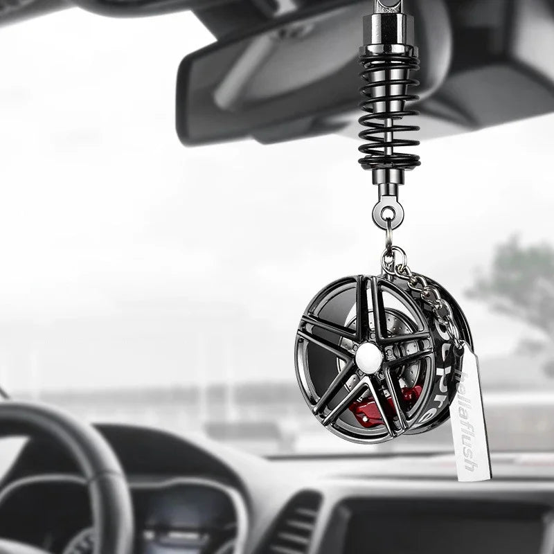 A black alloy wheel hub brake disc pendant hangs from a chain and is positioned in front of a car's rearview mirror.