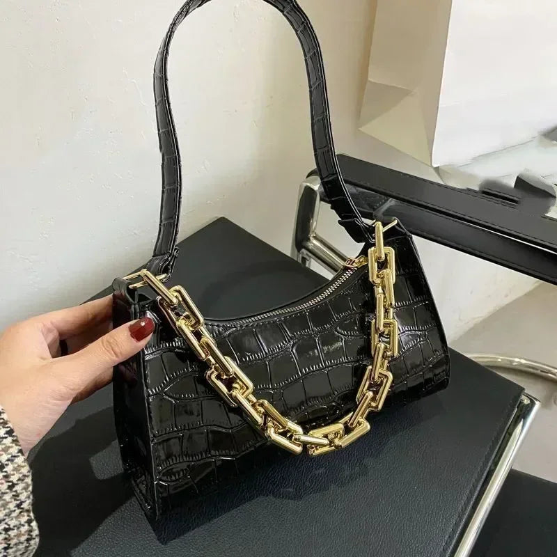 A black crocodile print shoulder bag with a gold chain strap is held by a hand on a dark surface.