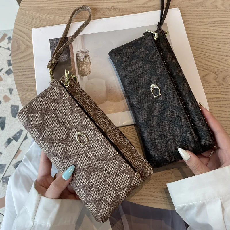 A hand holds a brown monogrammed zipper wallet with gold hardware against a white background.