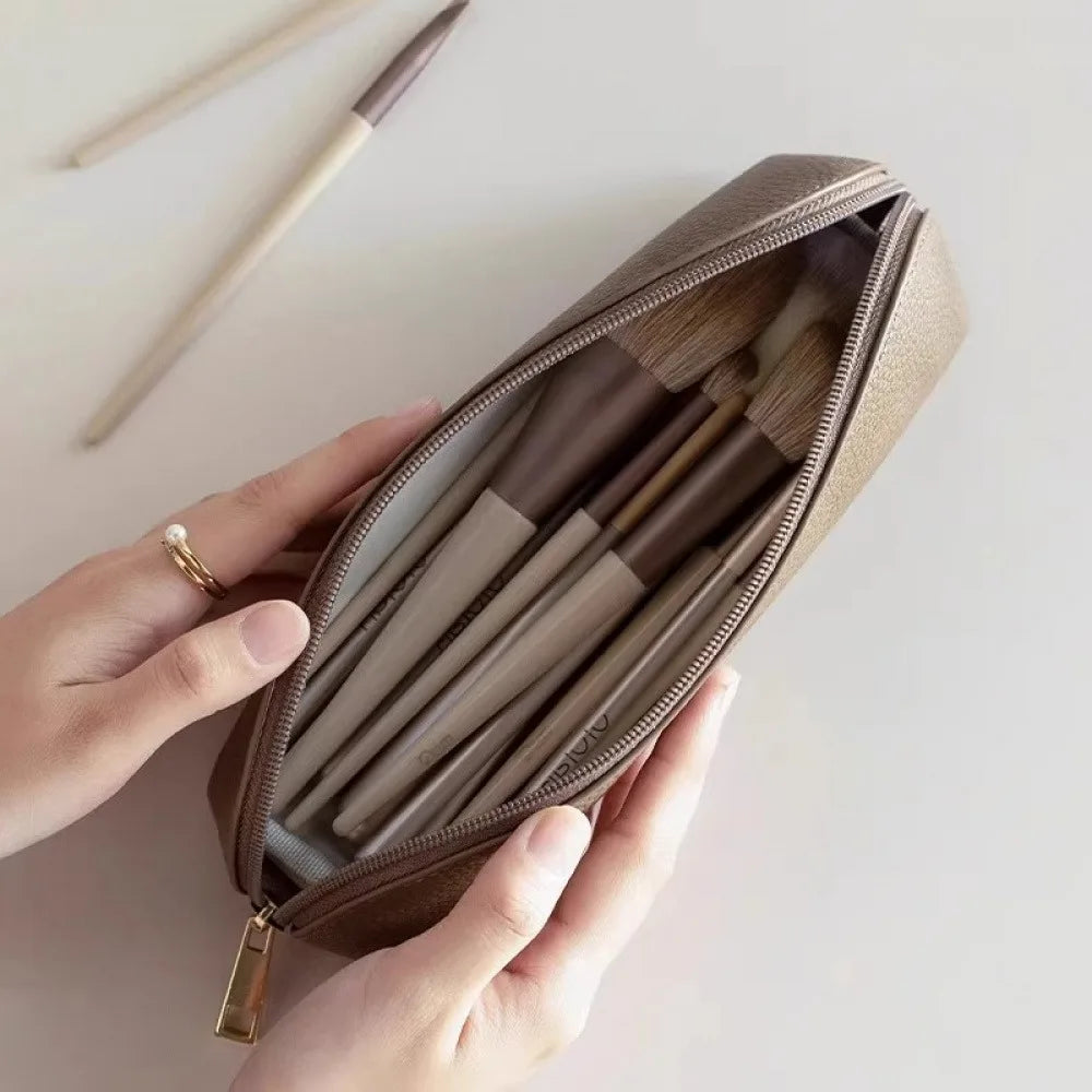 A hand holds an open brown makeup pouch revealing several pencils and cosmetic tools inside.