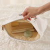 A hand is opening a white fabric storage pouch with a zipper to reveal a beige interior holding makeup brushes.