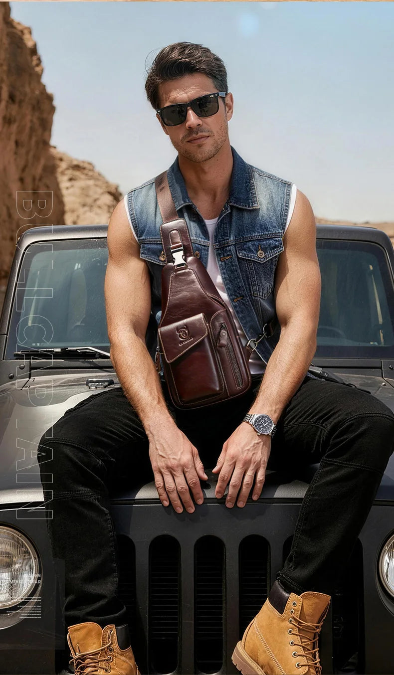 A man wearing a denim shirt sits on the hood of a dark car with a brown leather sling backpack.