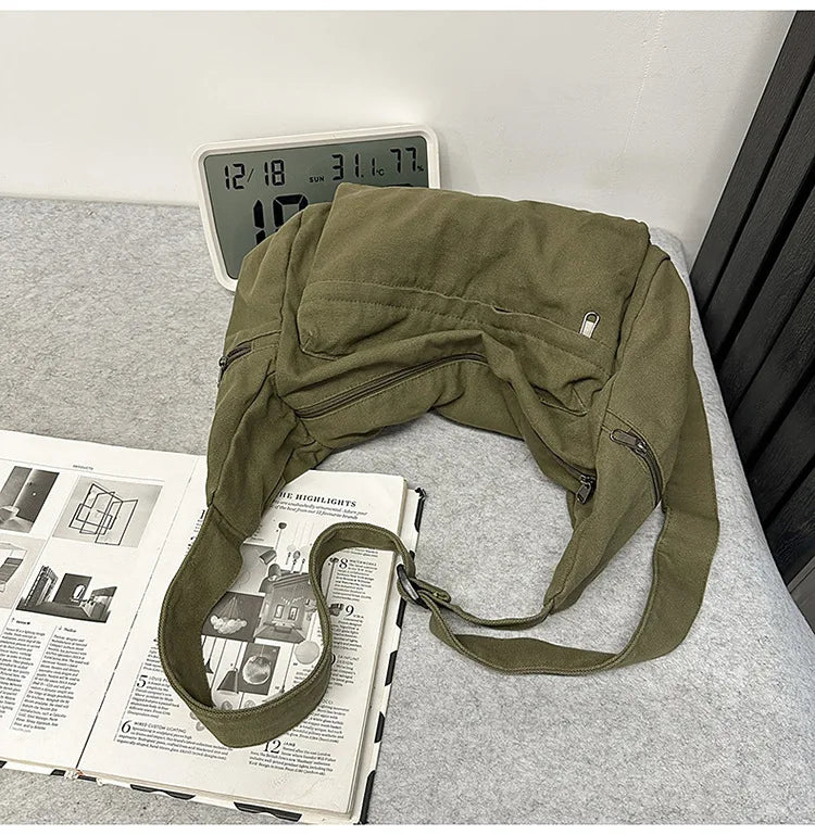 Olive green cotton canvas shoulder crossbody bag resting on a gray surface next to a digital clock and a book.