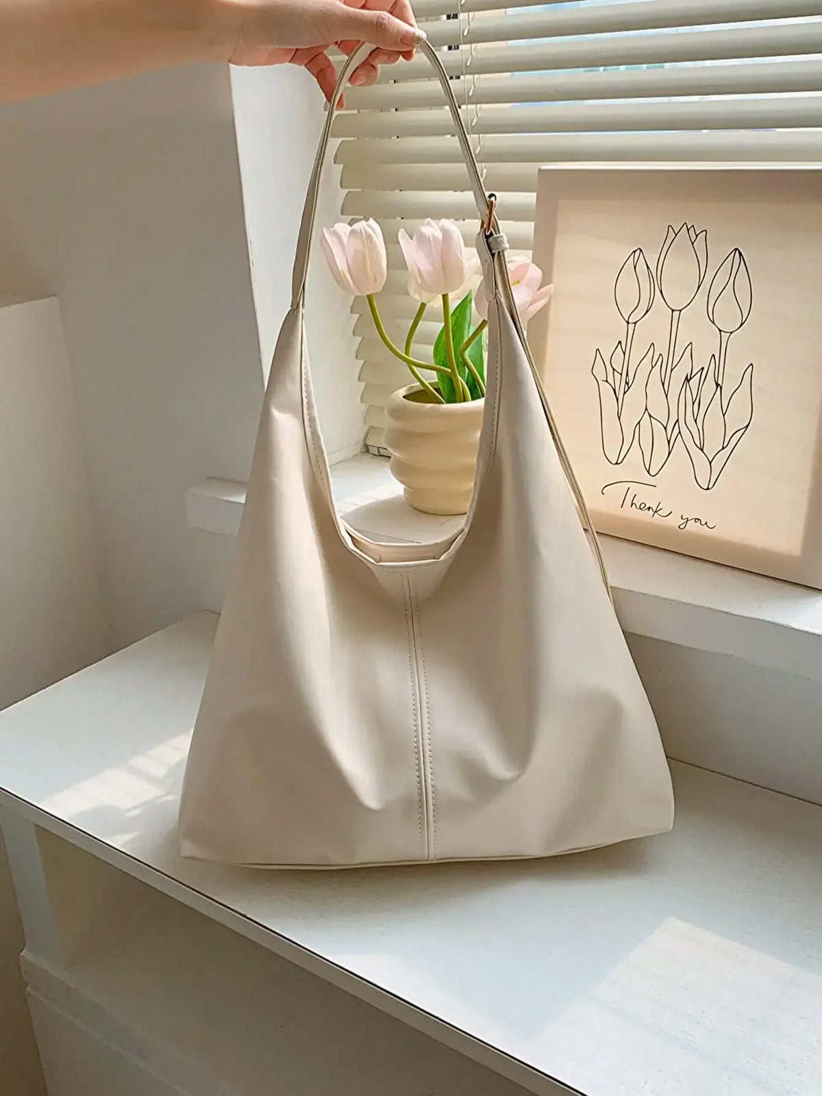 A beige PU leather slouchy hobo shoulder bag with a strap, placed on a white shelf next to a framed art print.