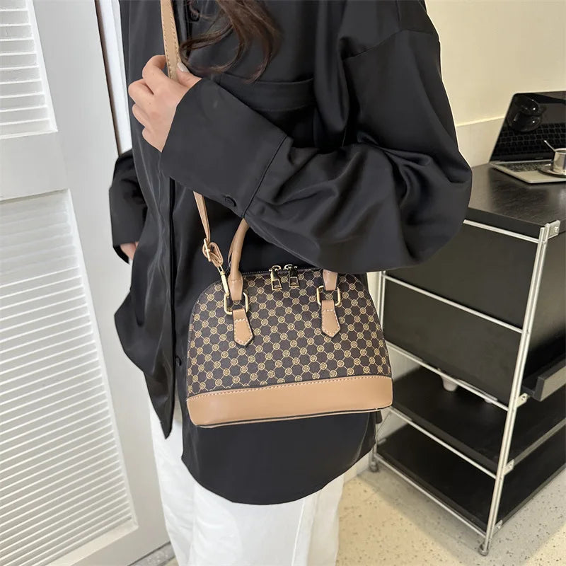 A woman holds a tan and black patterned fashion shell shoulder crossbody bag against a white background.
