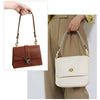 A brown leather handbag with a gold chain strap is shown being held by a hand against a beige skirt.
