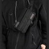 A person wearing a black hoodie holds a black chest bag across their body.