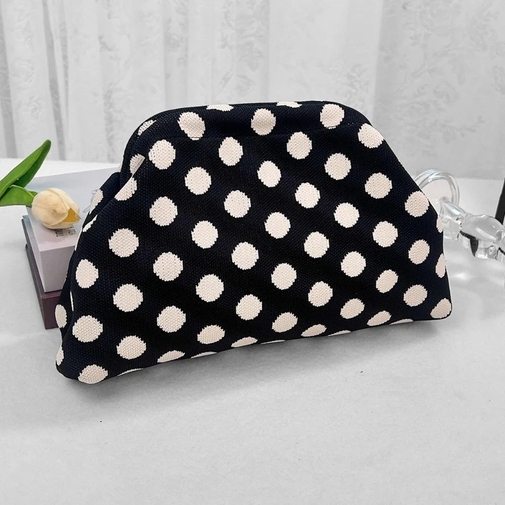 A black and white polka dot clutch bag is displayed on a white surface next to a light green flower.