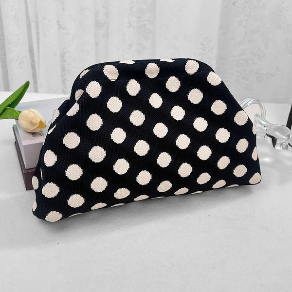 A black and white polka dot clutch bag is displayed on a white surface next to a light green flower.