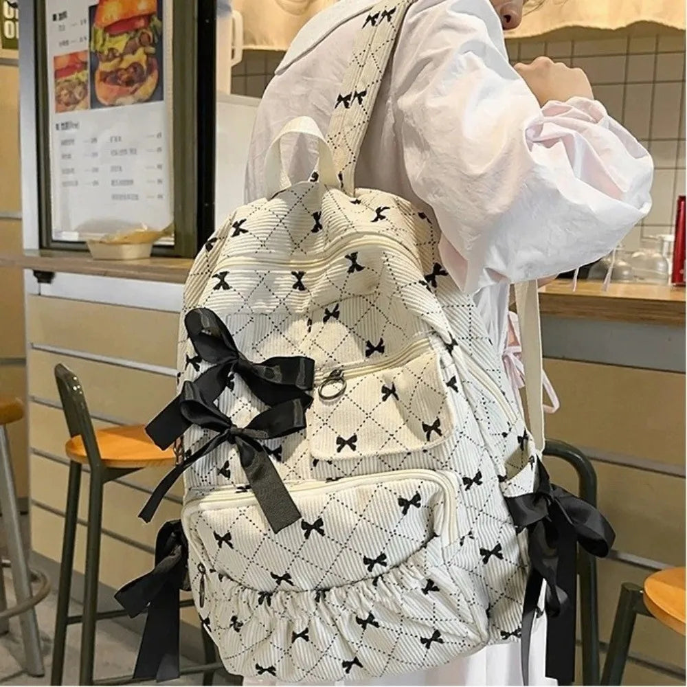 A woman holds a white corduroy bow print backpack with black ribbon details in a store setting.