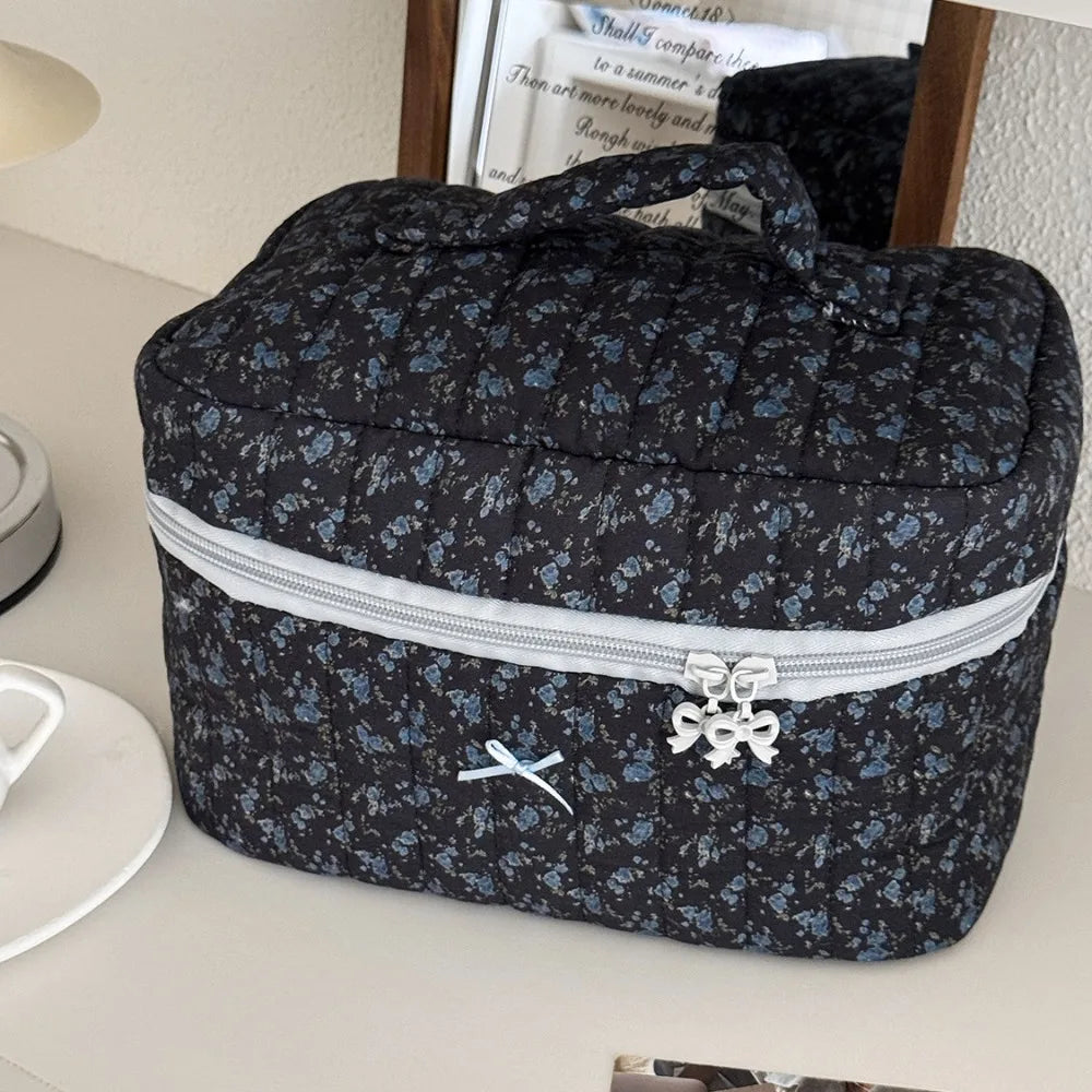 A dark floral patterned quilted cosmetic bag with a silver zipper and bow on a white surface.