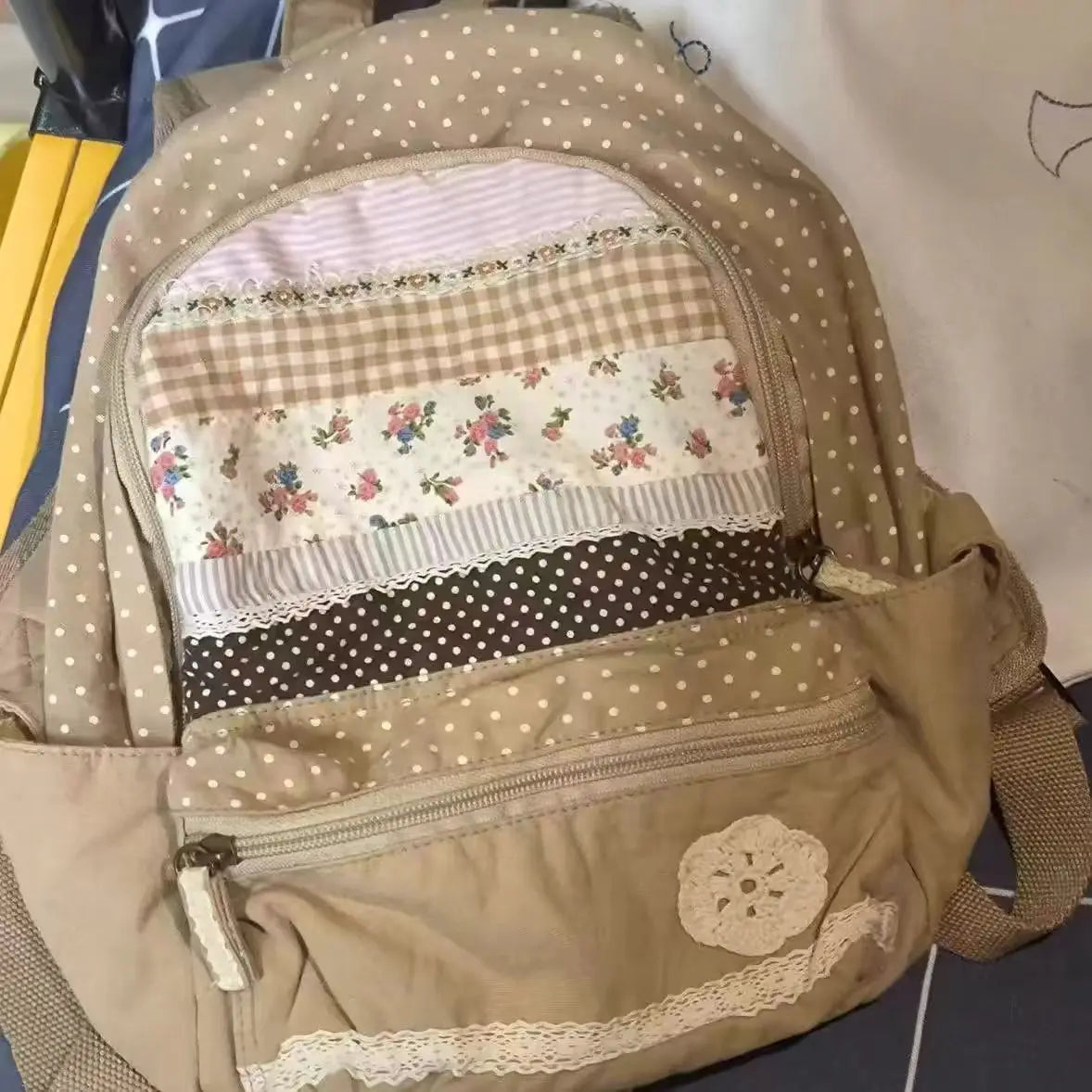 A beige backpack featuring layered fabric panels with floral and checkered patterns.