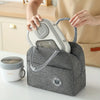A person is holding a gray insulated lunch bag containing a white food jar and a light gray electric food processor.