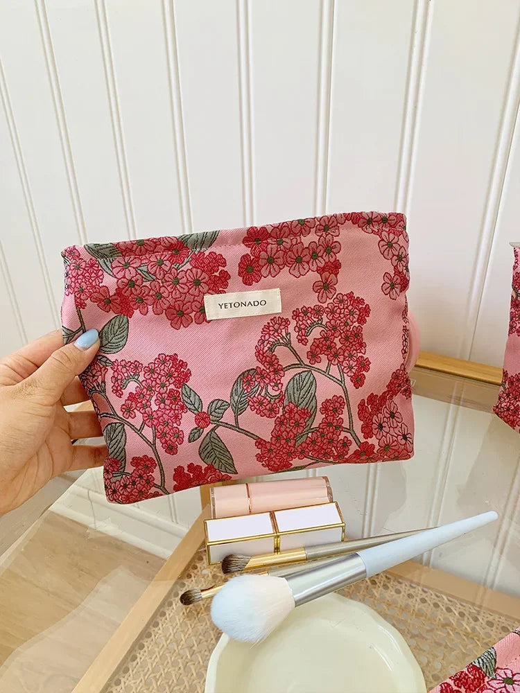 A pink cosmetic pouch with a floral pattern is held against a white paneled background.