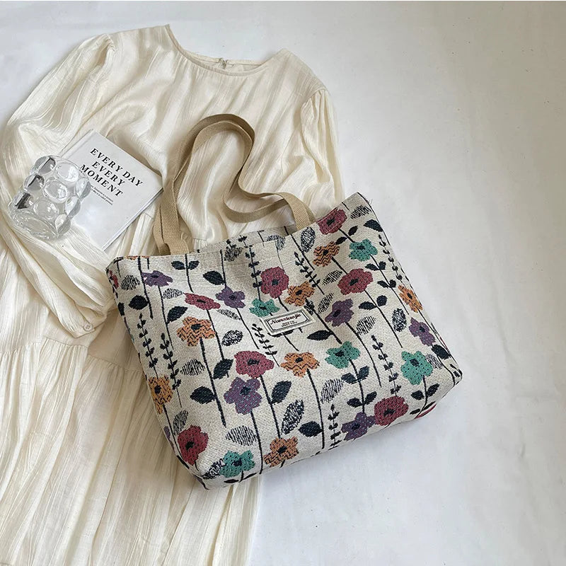 A beige bohemian corduroy tote bag with floral embroidery rests next to a white linen dress and a small glass charm.