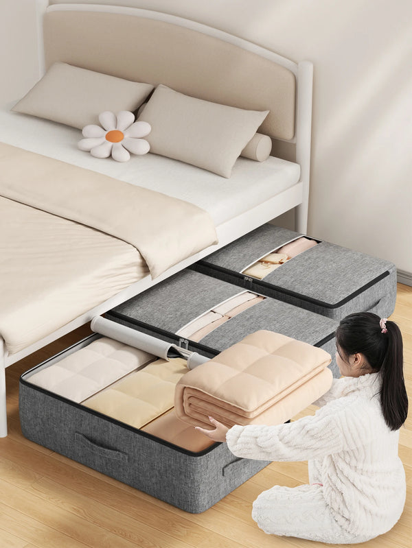 A woman is folding an extra-large foldable underbed storage bag with reinforced handles onto a bed in a bright room.