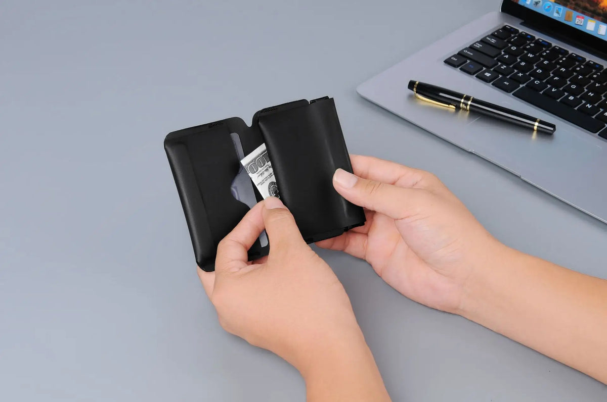 A hand holds a black magnetic foldable ID card holder and coin purse next to a silver card on a gray surface.