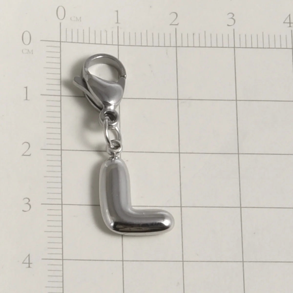 Silver stainless steel alphabet letter charm with a lobster clasp placed on a ruler.