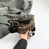 Eine Hand hält eine schwarz-leopardmusterige Crossbody-Bauchtasche mit goldfarbenen Verzierungen vor einem grauen Sakkojacke.