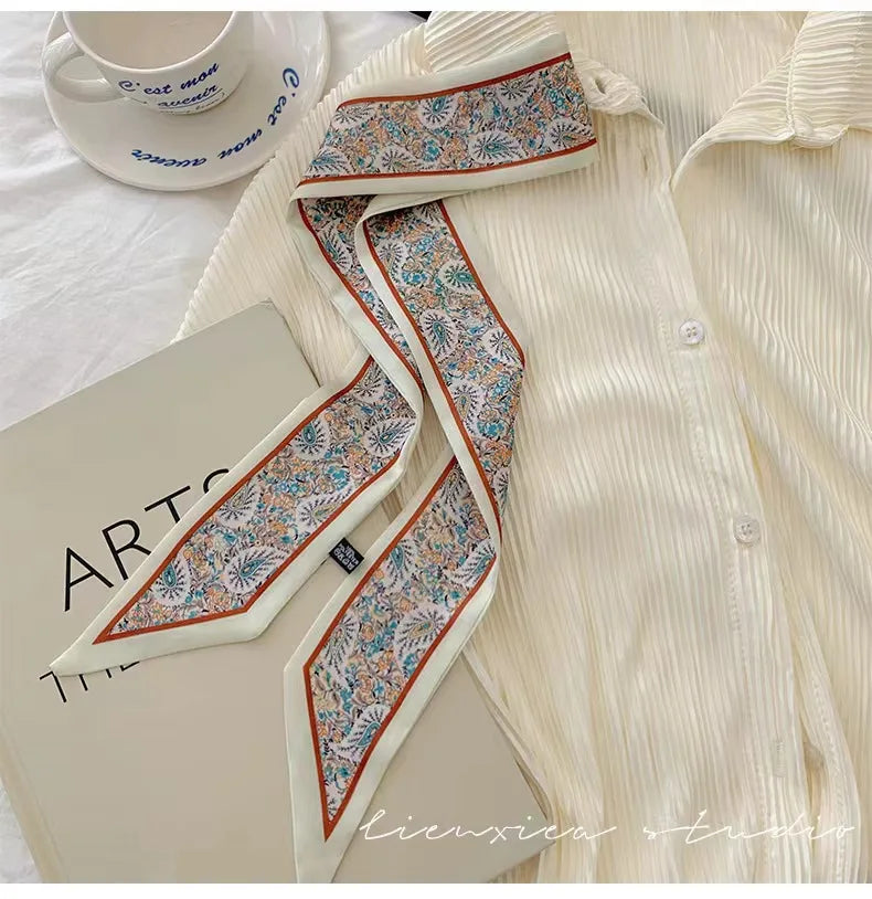 Chemise de couleur crème avec un foulard de soie à motif floral autour du cou, présentée à côté d'un carnet.