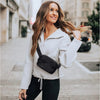 A woman wearing a white zip-up jacket and black pants is walking down a city street, carrying a black crossbody bag.