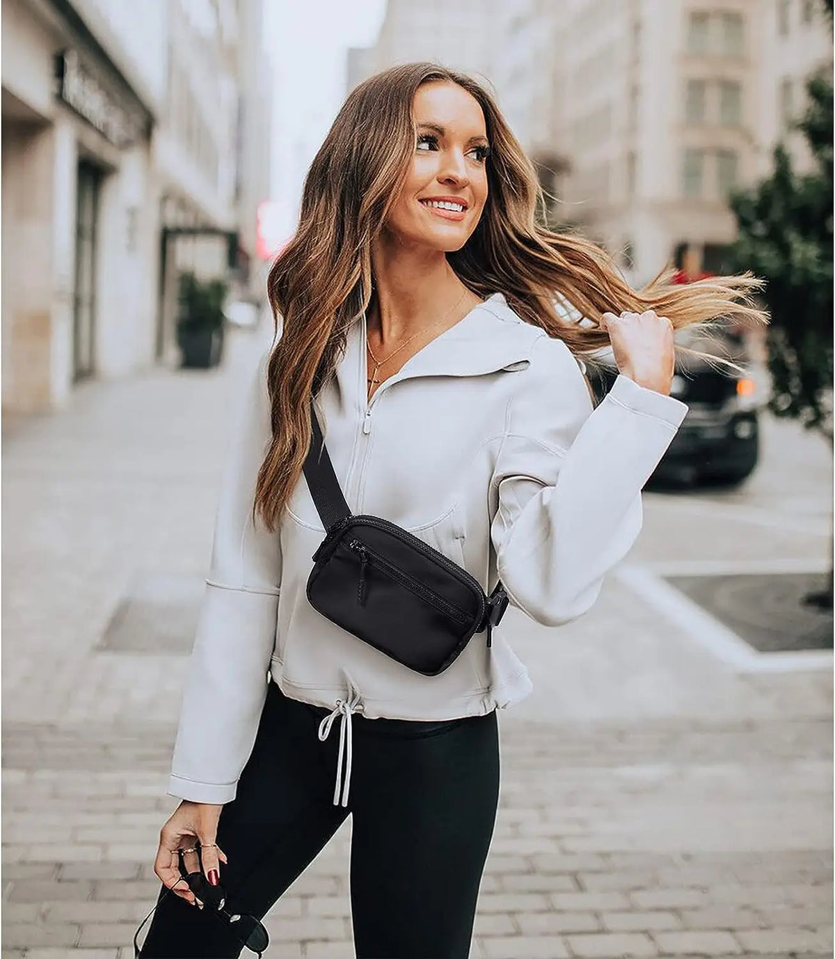 A woman wearing a white zip-up jacket and black pants is walking down a city street, carrying a black crossbody bag.
