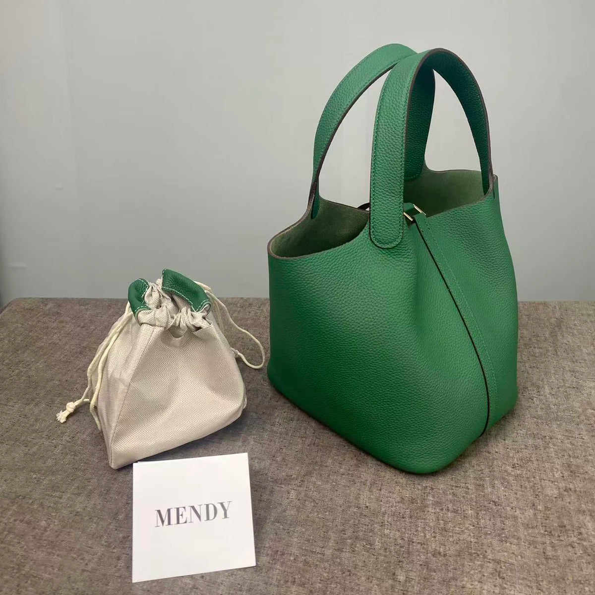 A deep green leather bucket bag next to a light beige fabric pouch and a card labeled MENDY.