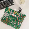 A green cosmetic pouch with floral embroidery rests on a white desk next to a laptop and makeup brushes.