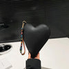 Cartera de monedas de PU con forma de corazón y color negro, sostenida contra un fondo oscuro.