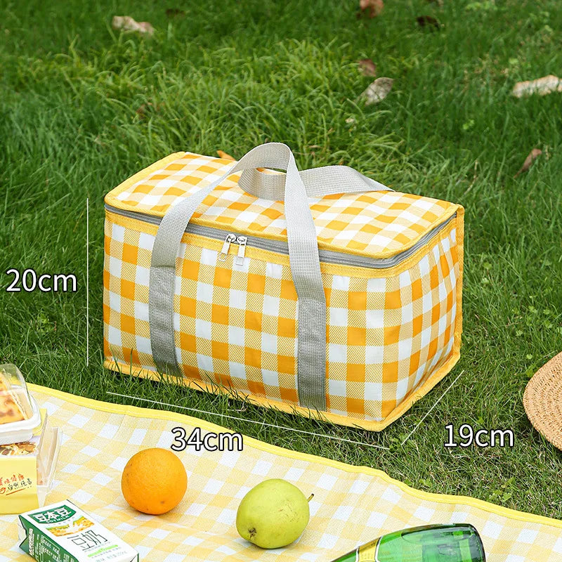 A yellow and white checkered insulated picnic tote bag lying on green grass with citrus fruits.