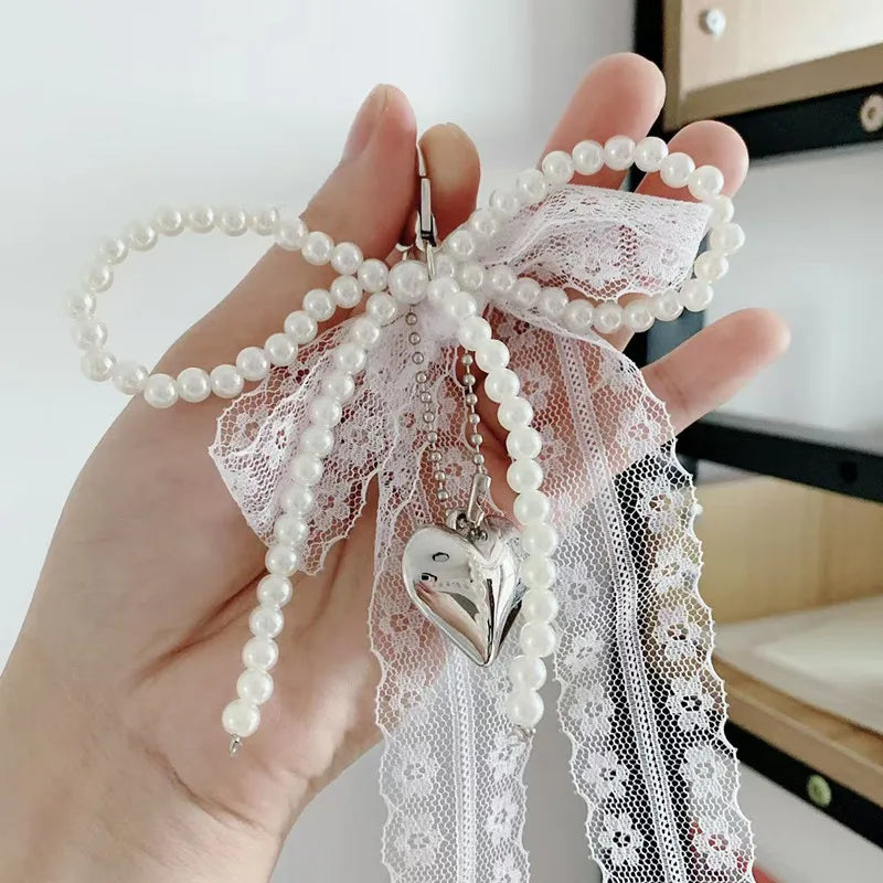 A hand holds a white pearl bow keychain featuring a heart-shaped pendant and delicate lace.