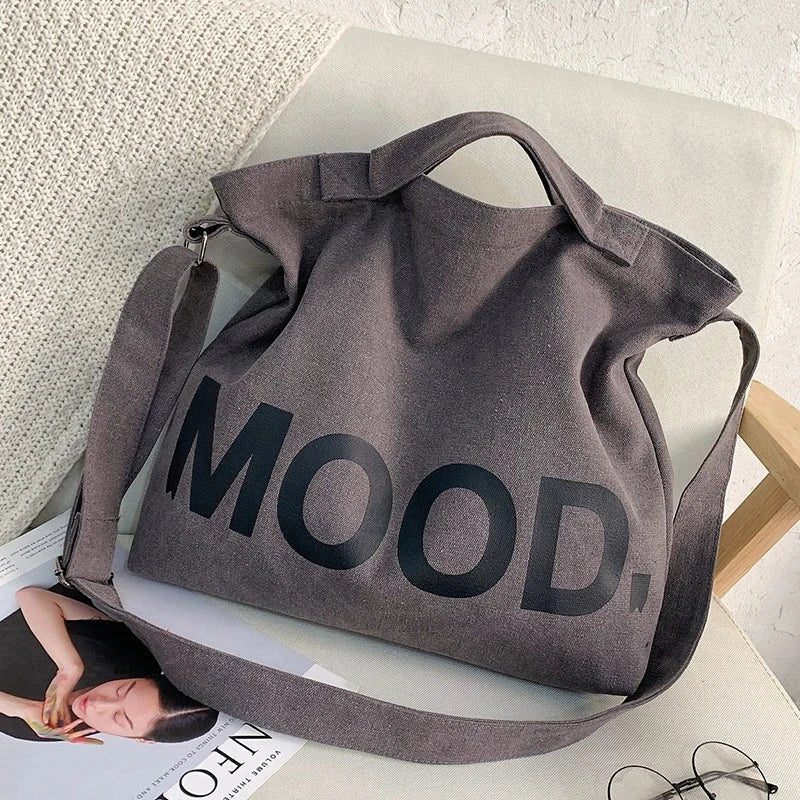 A gray canvas tote bag featuring the word MOOD in large black lettering, resting on a white surface.