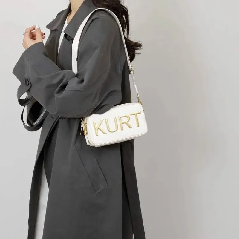 A woman holds a white leather tote bag with gold lettering against a gray coat.