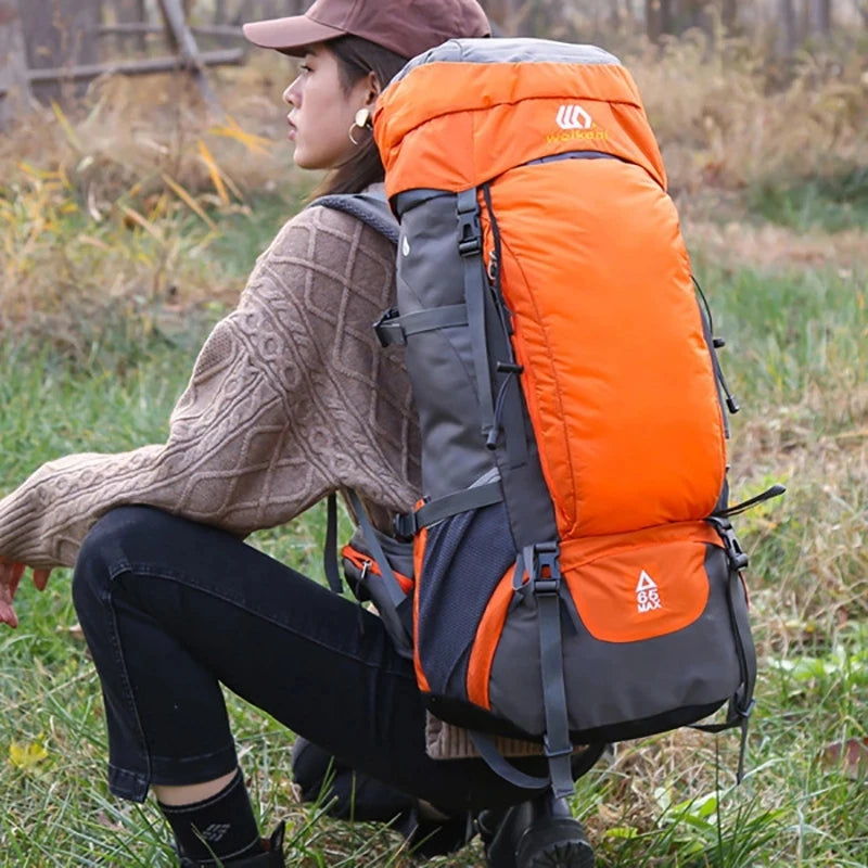 Une femme portant un pull marron et un pantalon noir est assise dehors avec un sac à dos d'escalade imperméable orange et gri