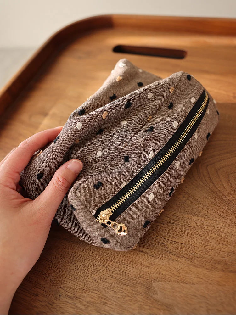 Une main tient une pochette de rangement cosmétique en tissu gris à pois noirs et une fermeture éclair dorée sur une surface 