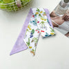 Un foulard pour les cheveux en soie violet avec un motif floral blanc, exposé à côté d'une bouteille en verre.