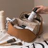 A hand is opening a tan cosmetic storage bag to reveal several makeup products and brushes.