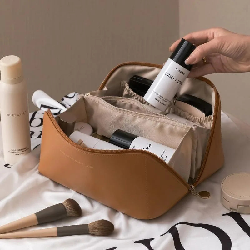A hand is opening a tan cosmetic storage bag to reveal several makeup products and brushes.