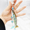A hand holds a silver fish-themed acrylic keychain charm with blue accents.
