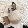 A beige soft leather crossbody bag with a zipper and adjustable strap resting on white bedding next to a smartphone.
