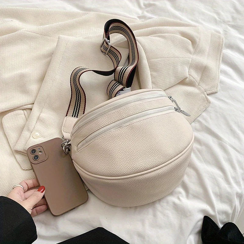 A beige soft leather crossbody bag with a zipper and adjustable strap resting on white bedding next to a smartphone.