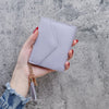 A hand holds a light purple PU leather coin clutch wallet with a gold chain tassel against a gray textured background.