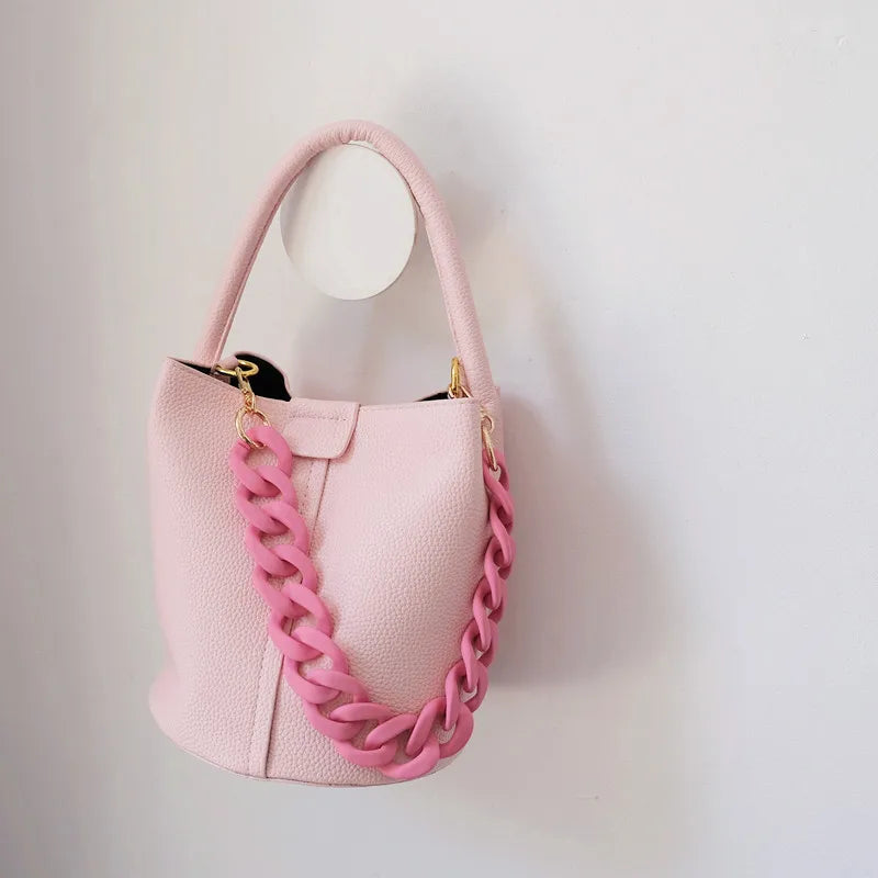 Sac à main en chaîne en résine rose mat, accroché contre un mur blanc.