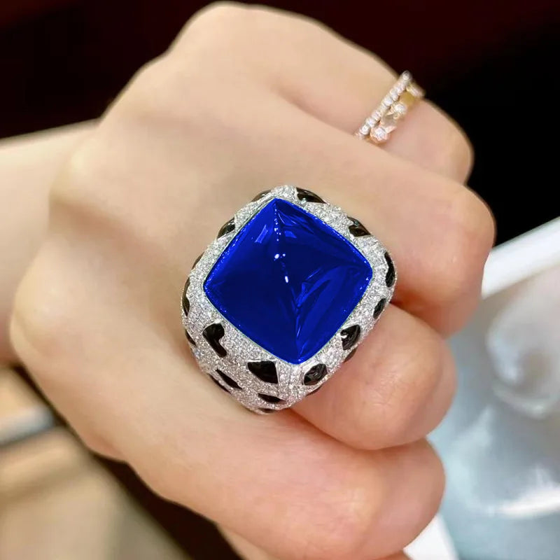 A hand is holding a luxury leopard crystal statement ring featuring a large blue gemstone set in a silver-like halo.